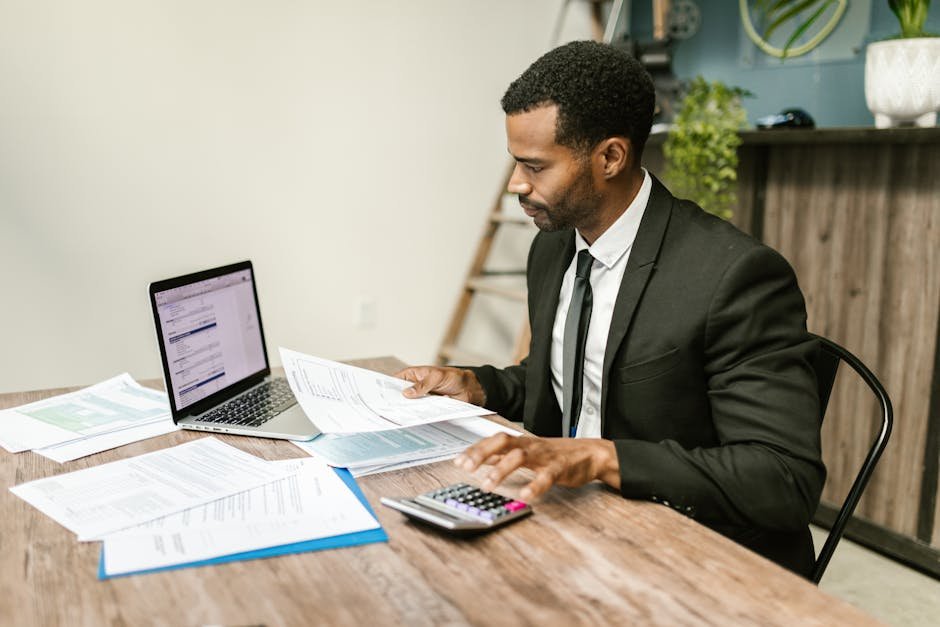 A businessman in a suit working on documents and using a laptop at an office desk.